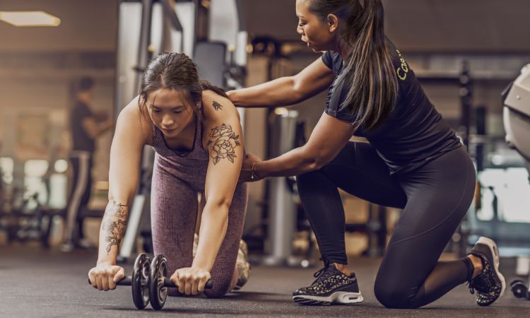 Personal trainer a orientar uma cliente num exercício com roda abdominal no ginásio, como se formar em personal trainer.